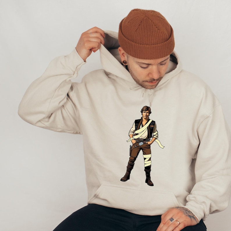 May include: A beige hoodie featuring a graphic of a character from a science fiction film. The character is wearing a brown vest, pants, and boots, with a white sash and a holster. The person in the image is wearing a brown beanie.