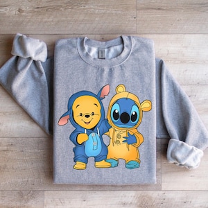 May include: Gray sweatshirt with a cartoon image of Stitch and Winnie the Pooh wearing matching outfits. Stitch is wearing a yellow raincoat and Winnie the Pooh is wearing a blue raincoat.