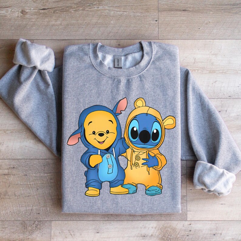 May include: Gray sweatshirt with a cartoon image of Stitch and Winnie the Pooh wearing matching outfits. Stitch is wearing a yellow raincoat and Winnie the Pooh is wearing a blue raincoat.