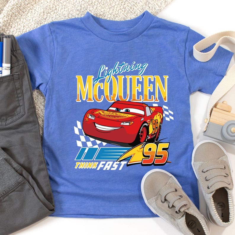Puede incluir: Una camiseta azul con Lightning McQueen, un coche de carreras rojo, y el texto "Lightning McQueen". La camiseta tambi&eacute;n tiene el texto "Think Fast" y el n&uacute;mero "95". Un pantal&oacute;n y zapatos grises est&aacute;n al lado de la camiseta.