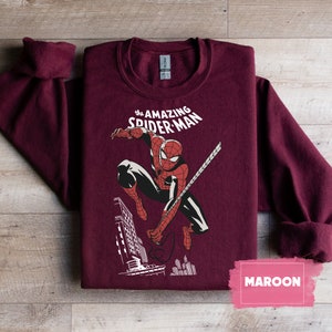 Pu&ograve; includere: Felpa color granata con un'immagine in stile fumetto vintage di Spider-Man che si dondola per la citt&agrave;. Il testo "the AMAZING SPIDER-MAN" &egrave; stampato sopra l'immagine.
