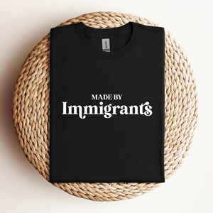 Może przedstawiać: Czarny T-shirt z białym napisem "MADE BY Immigrants".