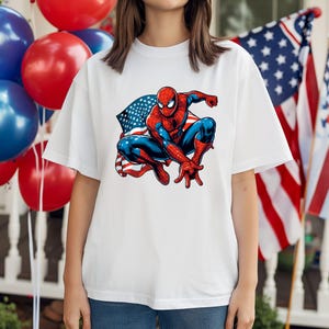 May include: White t-shirt featuring a graphic of Spider-Man in a crouching pose, set against an American flag backdrop. The superhero is depicted in his classic red and blue suit. Red, white, and blue balloons are visible in the background.
