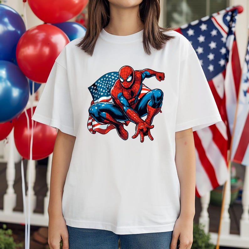 May include: White t-shirt featuring a graphic of Spider-Man in a crouching pose, set against an American flag backdrop. The superhero is depicted in his classic red and blue suit. Red, white, and blue balloons are visible in the background.