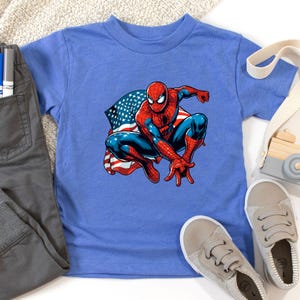 May include: A blue t-shirt featuring a graphic of Spider-Man in a crouching pose with an American flag design. The superhero is depicted in his classic red and blue suit. Gray pants and shoes are also visible.
