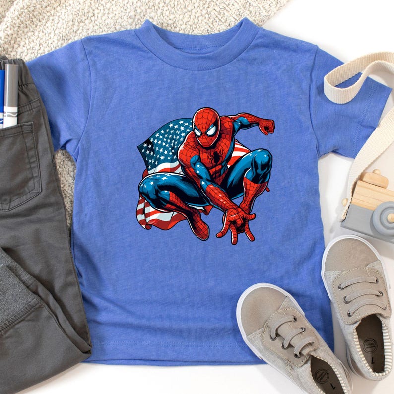 May include: A blue t-shirt featuring a graphic of Spider-Man in a crouching pose with an American flag design. The superhero is depicted in his classic red and blue suit. Gray pants and shoes are also visible.