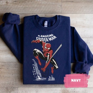 Pu&ograve; includere: Una felpa blu navy con una grafica di Spider-Man che si dondola attraverso la citt&agrave;. Il testo "the AMAZING SPIDER-MAN" &egrave; stampato sopra l'immagine.