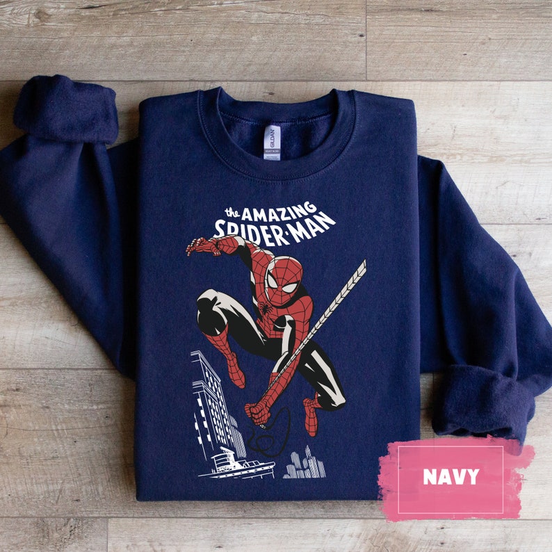 Pu&ograve; includere: Una felpa blu navy con una grafica di Spider-Man che si dondola attraverso la citt&agrave;. Il testo "the AMAZING SPIDER-MAN" &egrave; stampato sopra l'immagine.