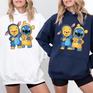 May include: Two white and navy blue hoodies with a graphic print of Winnie the Pooh and Stitch wearing matching outfits. The characters are in a cartoon style and are smiling.