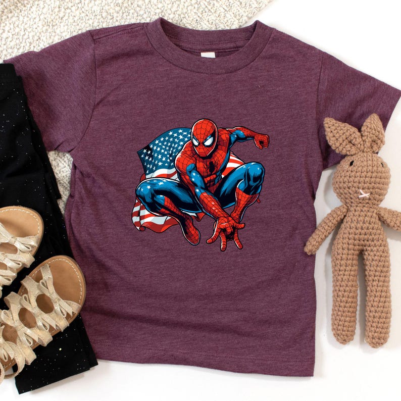 May include: A maroon t-shirt featuring a graphic of Spider-Man in a crouching pose with an American flag design. The superhero is depicted in his classic red and blue suit. The shirt is surrounded by black pants, sandals, and a stuffed bunny.