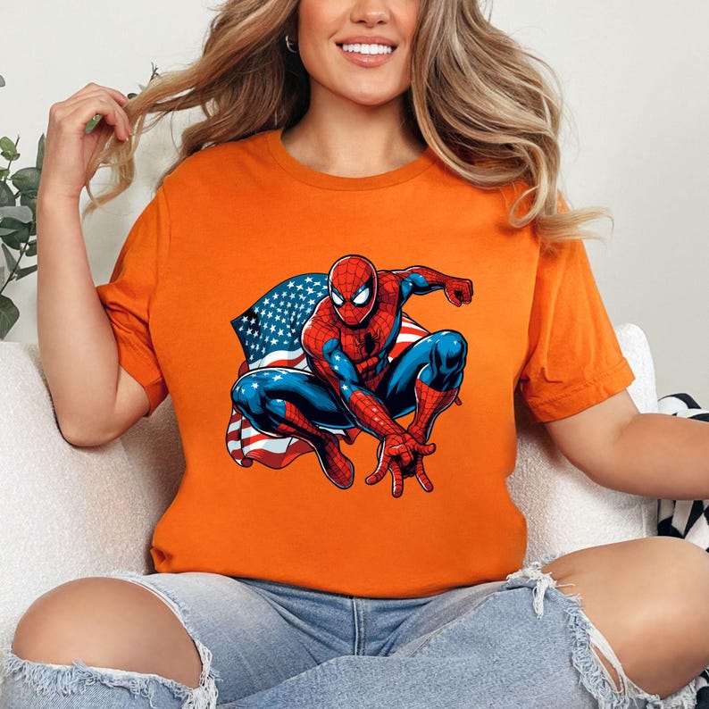 May include: Orange t-shirt featuring a graphic of Spider-Man with an American flag design. The superhero is depicted in his classic red and blue suit, with the flag draped behind him. The shirt is a crew neck style.