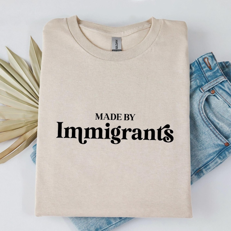 Może przedstawiać: Beżowa koszulka z nadrukiem "MADE BY Immigrants" w kolorze czarnym.