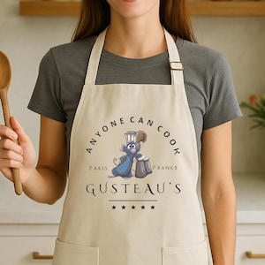May include: Beige apron with the text "ANYONE CAN COOK" and "GUSTEAU'S" in black. A cartoon rat wearing a chef's hat is featured in the center. The apron has a pocket and adjustable neck strap. The apron is worn by a person.