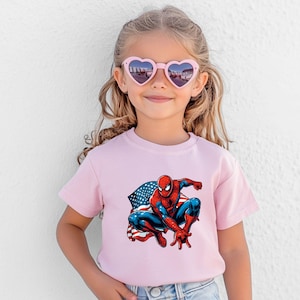 May include: A light pink t-shirt featuring a graphic of Spider-Man in a crouching pose with the American flag. The superhero is depicted in his classic red and blue suit. The girl is wearing pink heart-shaped sunglasses.