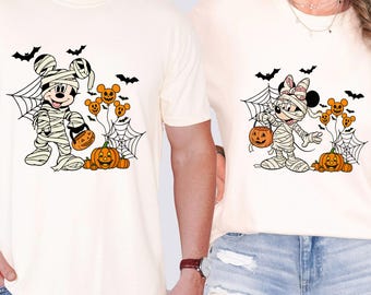 May include: Two cream-colored t-shirts featuring Halloween-themed cartoon characters. Mickey Mouse and Minnie Mouse are dressed as mummies, holding jack-o'-lanterns. Bats, spiderwebs, and pumpkin balloons complete the festive design.