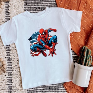 May include: White t-shirt featuring a colorful illustration of Spider-Man in a crouching pose. The superhero is depicted in his classic red and blue suit, with an American flag design in the background.