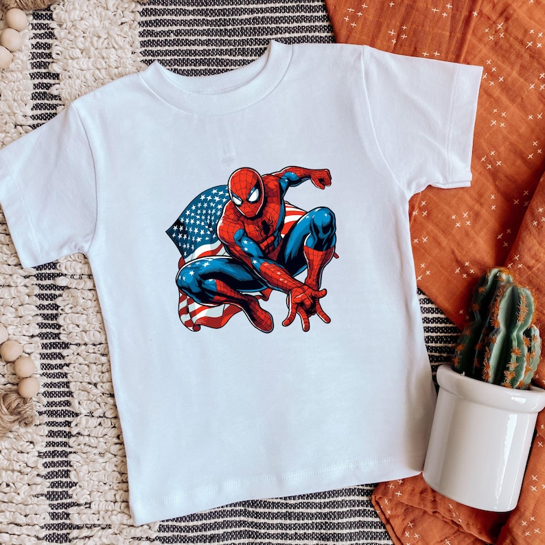 May include: White t-shirt featuring a colorful illustration of Spider-Man in a crouching pose. The superhero is depicted in his classic red and blue suit, with an American flag design in the background.