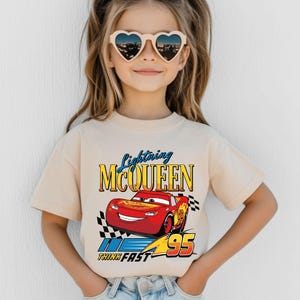 Puede incluir: Una camiseta beige con un dise&ntilde;o de Lightning McQueen, un coche de carreras rojo de la pel&iacute;cula Cars. El dise&ntilde;o incluye el texto "Lightning McQueen", "95" y "Think Fast". La camiseta la lleva un ni&ntilde;o con gafas de sol en forma de coraz&oacute;n.