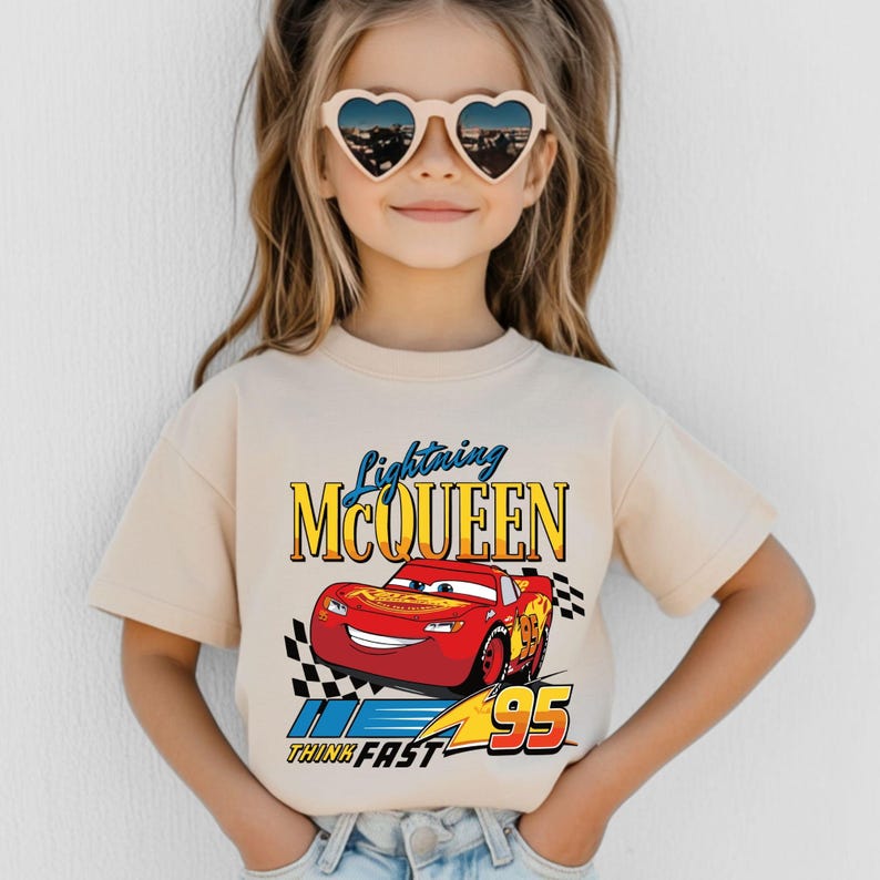 Puede incluir: Una camiseta beige con un dise&ntilde;o de Lightning McQueen, un coche de carreras rojo de la pel&iacute;cula Cars. El dise&ntilde;o incluye el texto "Lightning McQueen", "95" y "Think Fast". La camiseta la lleva un ni&ntilde;o con gafas de sol en forma de coraz&oacute;n.