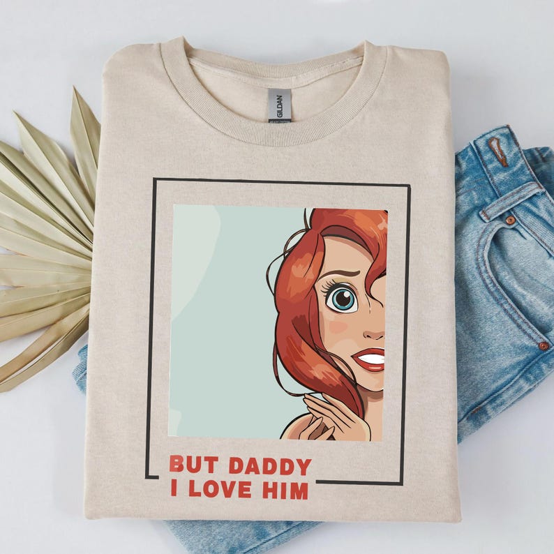 May include: A beige t-shirt featuring a cartoon image of a woman with red hair and the text "But Daddy I Love Him" printed on the front.
