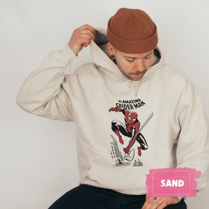 Pu&ograve; includere: Una felpa con cappuccio beige con una stampa grafica di Spiderman che oscilla su una ragnatela. Il testo "The Amazing Spider-Man" &egrave; stampato sopra l'immagine. La felpa ha un cappuccio con coulisse e una tasca a marsupio. Il modello indossa un berretto marrone.