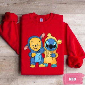 May include: Red sweatshirt with a cartoon image of Winnie the Pooh and Stitch wearing matching blue and yellow outfits. The text "RED" is visible in the bottom right corner of the image.