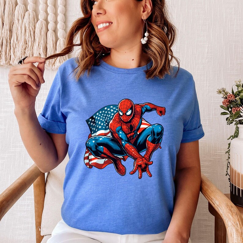 May include: A light blue t-shirt featuring a graphic of Spider-Man in a crouching pose with an American flag backdrop. The superhero is depicted in his classic red and blue suit. The shirt has short sleeves and a crew neck.
