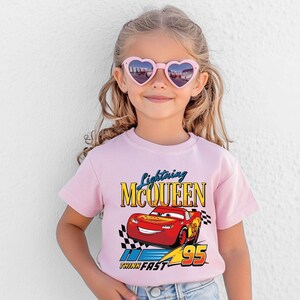 Puede incluir: Una camiseta rosa claro con un dise&ntilde;o de Rayo McQueen, el personaje de la pel&iacute;cula Cars. El dise&ntilde;o incluye el texto "Lightning McQueen", el n&uacute;mero "95" y la frase "Think Fast". La camiseta la lleva una persona joven con gafas de sol en forma de coraz&oacute;n.