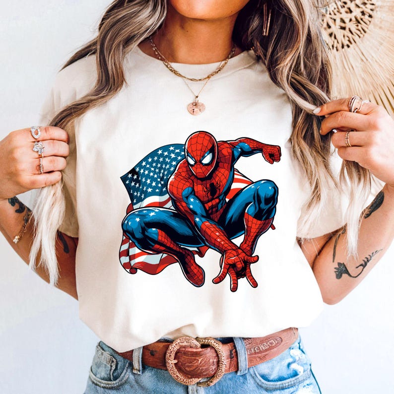 May include: Cream-colored t-shirt featuring a graphic of Spider-Man in a crouching pose, with the American flag as a backdrop. The superhero is depicted in his classic red and blue suit. The shirt has short sleeves and a crew neck.
