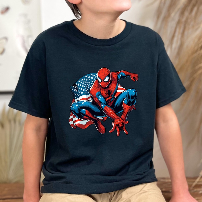 May include: Navy blue t-shirt featuring a graphic of Spider-Man in a crouching pose with the American flag as a backdrop. The superhero is depicted in his classic red and blue suit. The shirt is made of a soft, comfortable material.