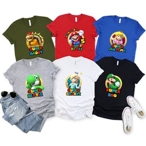 May include: Six t-shirts featuring different characters from the Super Mario Bros. video game franchise. The shirts are in various colors, including green, red, blue, gray, white, and black. Each shirt features a different character, including Mario, Luigi, Peach, Bowser, Yoshi, and Princess Daisy. The text on the shirts reads "Super Daddy", "Birthday Boy 5 Super James", "Super Mommy", "Super Bestie", "Super Granny", and "Super Bro".