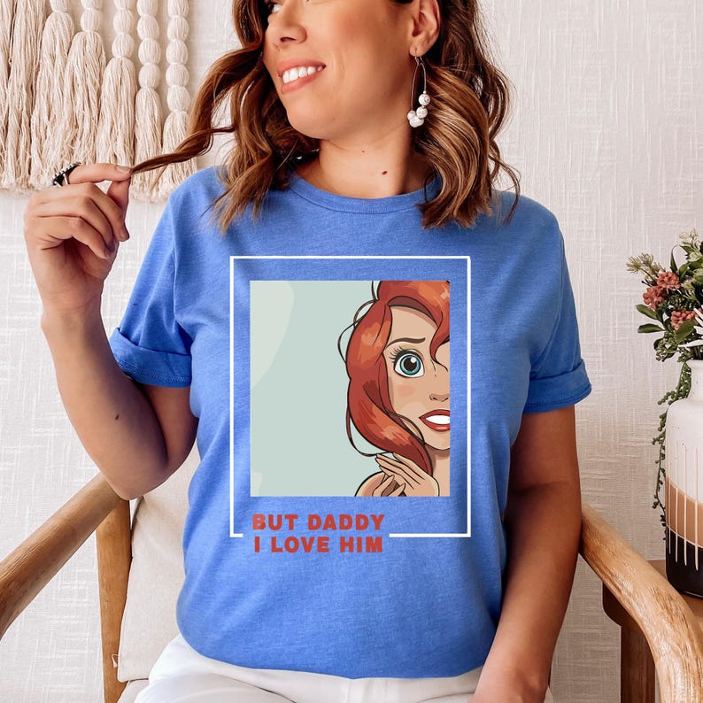 May include: A blue t-shirt featuring a cartoon image of a red-haired woman with the text "But Daddy I Love Him" printed on the front.
