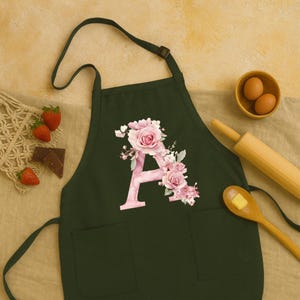 May include: Dark green apron with two pockets and a neck strap. The apron features a large pink letter "A" decorated with pink roses and floral accents. Strawberries, chocolate, eggs, and a rolling pin are in the background.