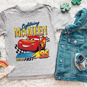 Puede incluir: Una camiseta gris de manga corta con Lightning McQueen de la pel&iacute;cula Cars. El gr&aacute;fico incluye el texto "Lightning McQueen", "Think Fast" y el n&uacute;mero "95". Una chaqueta vaquera est&aacute; al lado de la camiseta.