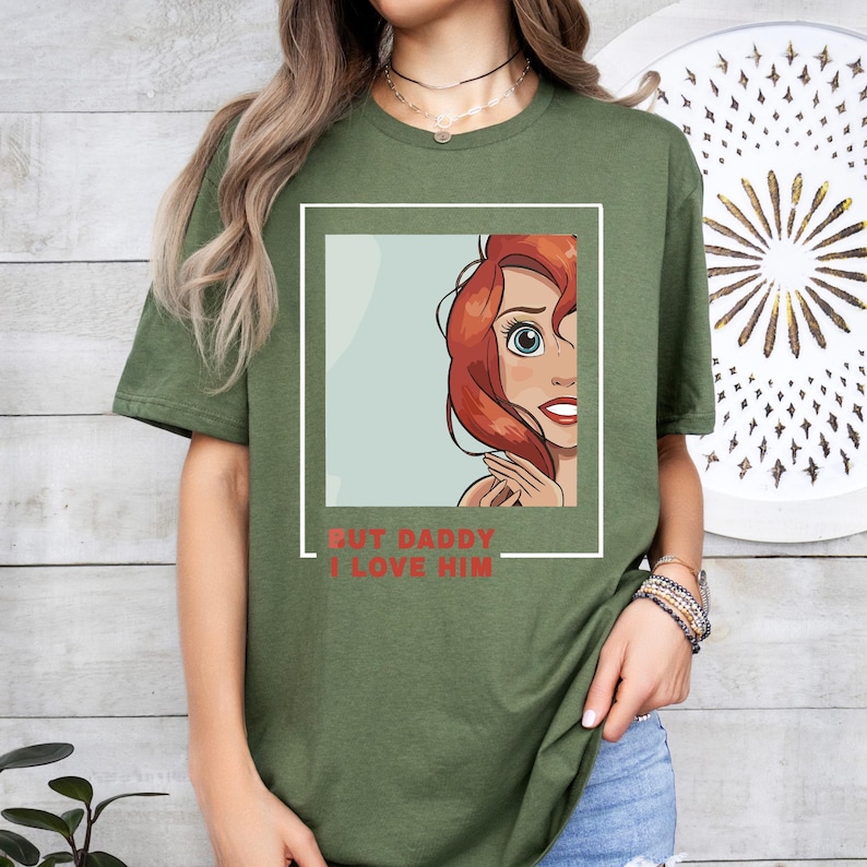 May include: Green t-shirt featuring a cartoon image of a woman with red hair. The text "But Daddy I Love Him" is printed on the shirt.