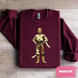 May include: Maroon sweatshirt featuring a gold-colored illustration of C-3PO from Star Wars. The character is depicted with gold metallic plating and wrapped with white bandages. The word "MAROON" is printed on a pink rectangle.