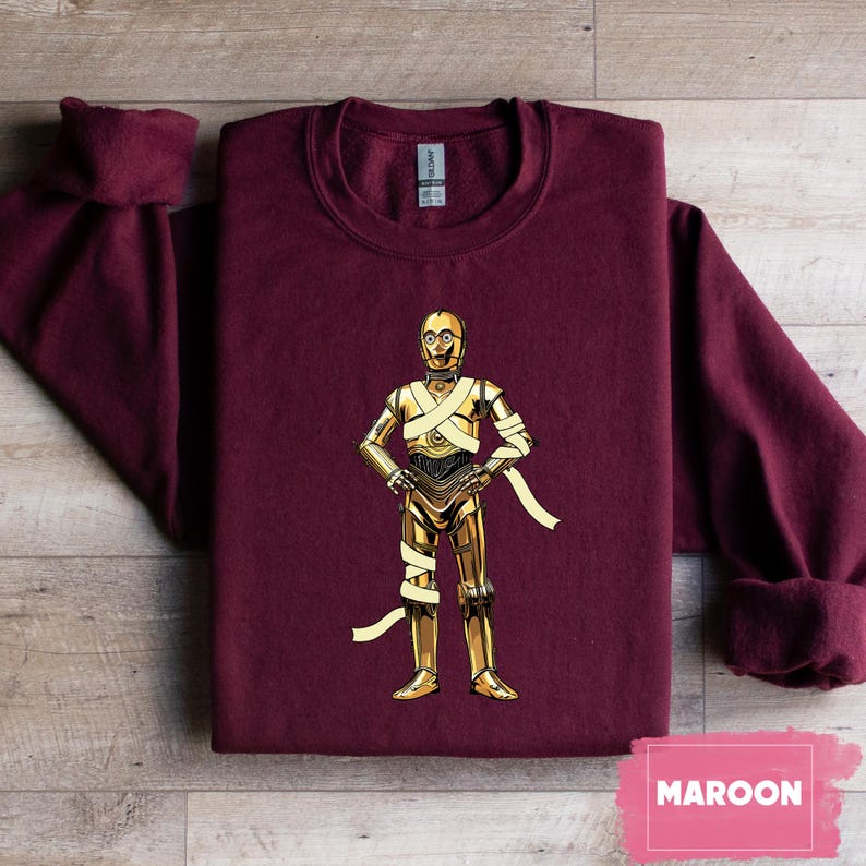May include: Maroon sweatshirt featuring a gold-colored illustration of C-3PO from Star Wars. The character is depicted with gold metallic plating and wrapped with white bandages. The word "MAROON" is printed on a pink rectangle.
