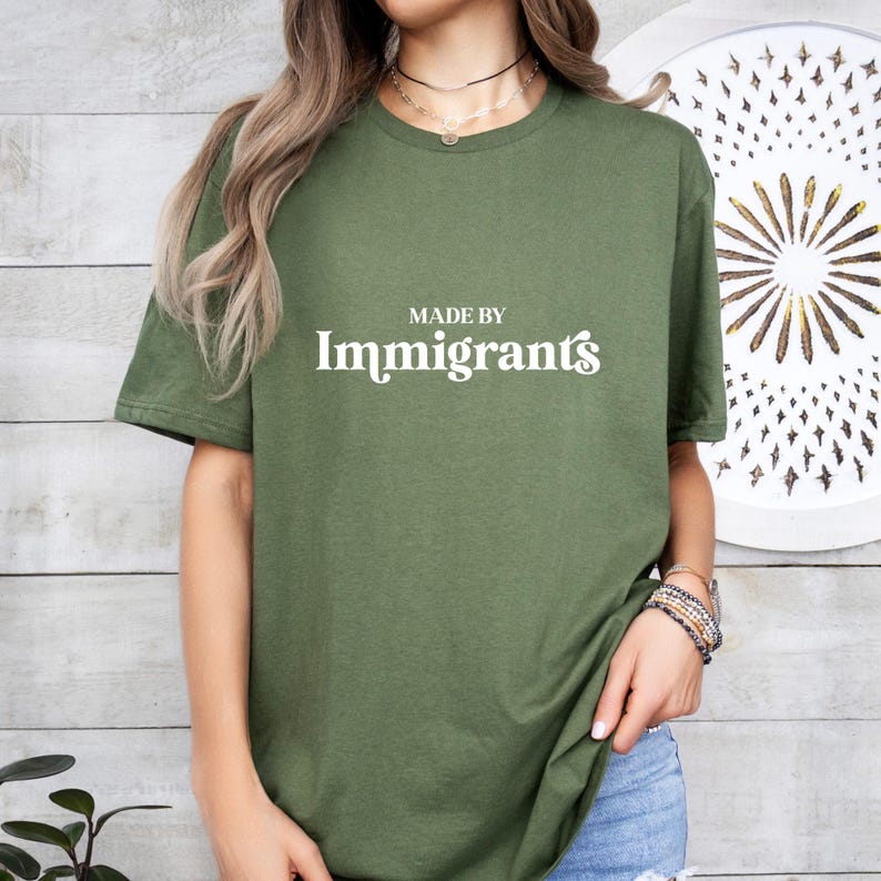 Może przedstawiać: Zielona koszulka z napisem "MADE BY Immigrants" w białym kolorze.