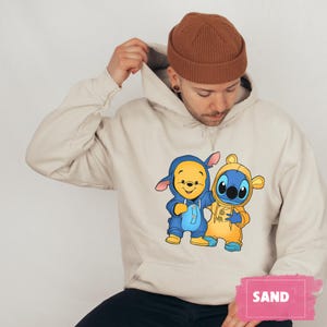 May include: A beige hoodie with a graphic print of Winnie the Pooh and Stitch wearing matching outfits. The hoodie has a drawstring hood and a kangaroo pocket.