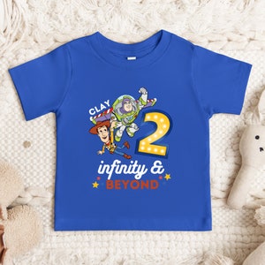Puede incluir: Una camiseta azul para ni&ntilde;os peque&ntilde;os con un gr&aacute;fico de Woody y Buzz Lightyear de Toy Story. La camiseta tiene el texto "CLAY" e "infinity & BEYOND". El n&uacute;mero "2" tambi&eacute;n est&aacute; en la camiseta, con bombillas alrededor.
