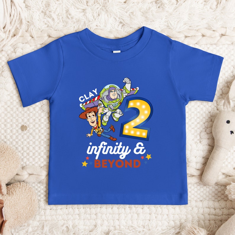 Puede incluir: Una camiseta azul para ni&ntilde;os peque&ntilde;os con un gr&aacute;fico de Woody y Buzz Lightyear de Toy Story. La camiseta tiene el texto "CLAY" e "infinity & BEYOND". El n&uacute;mero "2" tambi&eacute;n est&aacute; en la camiseta, con bombillas alrededor.