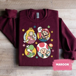 May include: Maroon sweatshirt with a graphic print of Mario, Luigi, Peach, and Yoshi. The characters are in circles with a yellow star background. The text "MAROON" is in a pink box at the bottom right corner of the image.