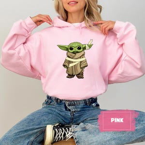 May include: A light pink hoodie featuring a graphic of a green alien character with large eyes, wearing a brown cloak and a bandage. The word "PINK" is displayed in a pink rectangle. The hoodie is worn by a person.
