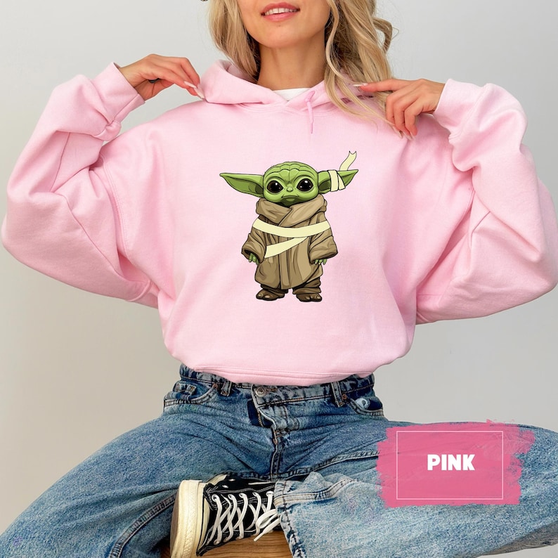 May include: A light pink hoodie featuring a graphic of a green alien character with large eyes, wearing a brown cloak and a bandage. The word "PINK" is displayed in a pink rectangle. The hoodie is worn by a person.