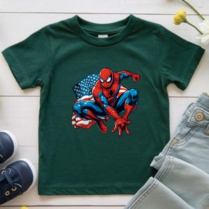 May include: A dark green t-shirt featuring a graphic of Spider-Man in a crouching pose with an American flag design in the background. The superhero is depicted in his classic red and blue suit.