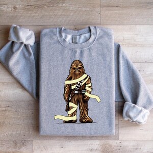 May include: A gray sweatshirt featuring a brown and tan graphic of a Wookiee-like character with a utility belt and wrapped in yellow bands. The sweatshirt has long sleeves and a crew neck.
