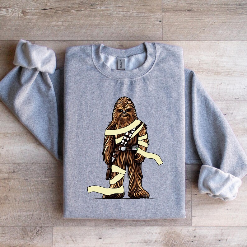 May include: A gray sweatshirt featuring a brown and tan graphic of a Wookiee-like character with a utility belt and wrapped in yellow bands. The sweatshirt has long sleeves and a crew neck.