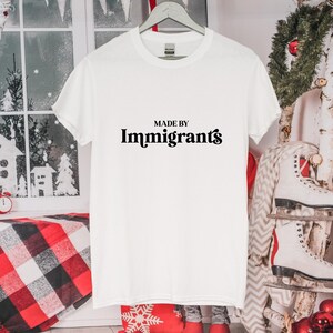 Może przedstawiać: Biała koszulka z czarnym napisem "MADE BY Immigrants" w stylizowanym kroju pisma.