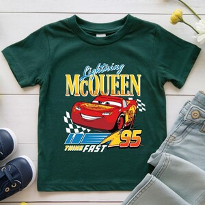 Puede incluir: Una camiseta verde oscuro con un dise&ntilde;o de Lightning McQueen, un coche de carreras rojo y el texto "Lightning McQueen". El dise&ntilde;o tambi&eacute;n incluye el n&uacute;mero "95" y las palabras "Think Fast".