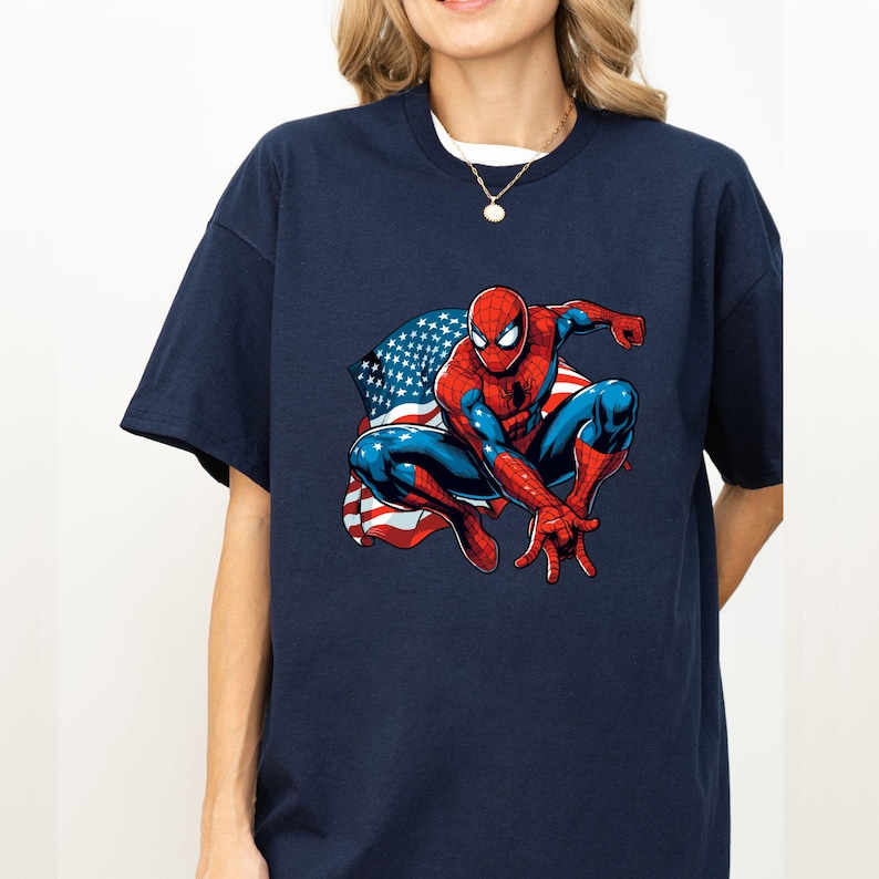 May include: Navy blue t-shirt featuring a graphic of Spider-Man in a crouching pose with an American flag design in the background. The superhero is depicted in his classic red and blue suit.